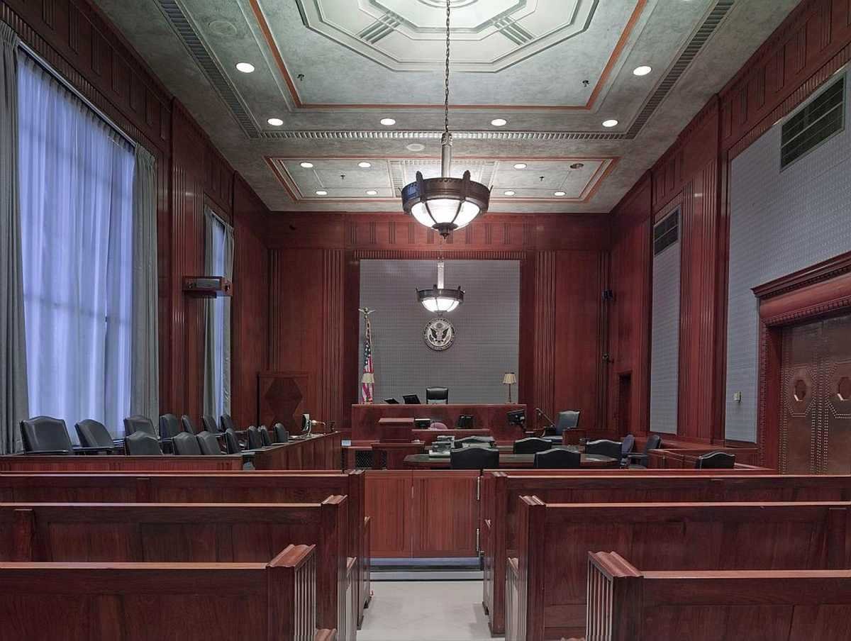 Salle d'audience aux boiseries rouges, bancs vides, plafond géométrique, luminaires suspendus